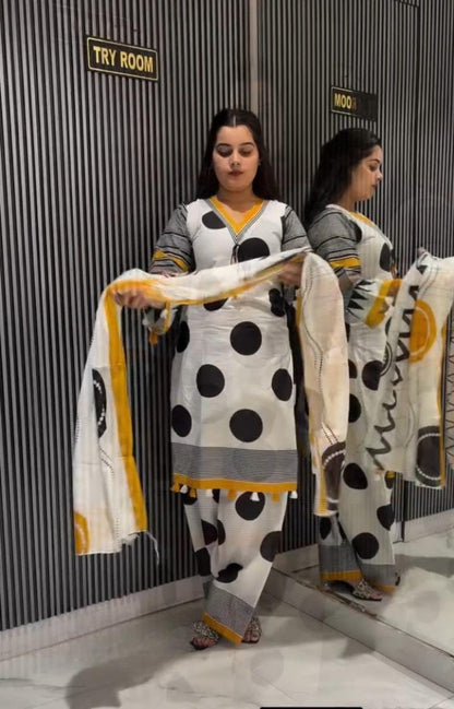 A woman stands in a fitting room wearing a white salwar suit with large black polka dots and gray sleeves, holding a matching dupatta with yellow and black accents. A mirror reflects her profile, and signs on the wall read 'TRY ROOM' and 'WOO'.